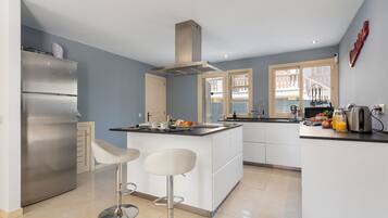 Een moderne keuken met een roestvrijstalen koelkast, witte barstoelen, een centraal eiland en een groot raam.
