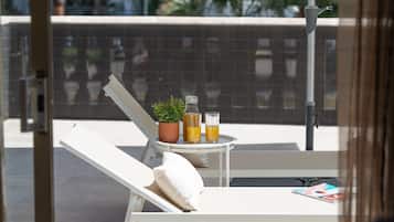 Een zonnig balkon met een witte lounge stoel, een tafeltje met een plant in een pot, twee glazen sinaasappelsap en een waterfles.