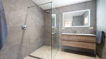 Een moderne badkamer met een douchecabine van glas, een grote spiegel met ingebouwde verlichting en een houten wastafelmeubel met wastafel.