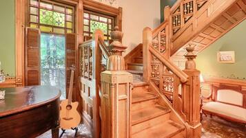 Una majestuosa escalera con detalles tallados, un piano y una guitarra en una habitación cálidamente iluminada, con ventanas amplias y una pared verde.