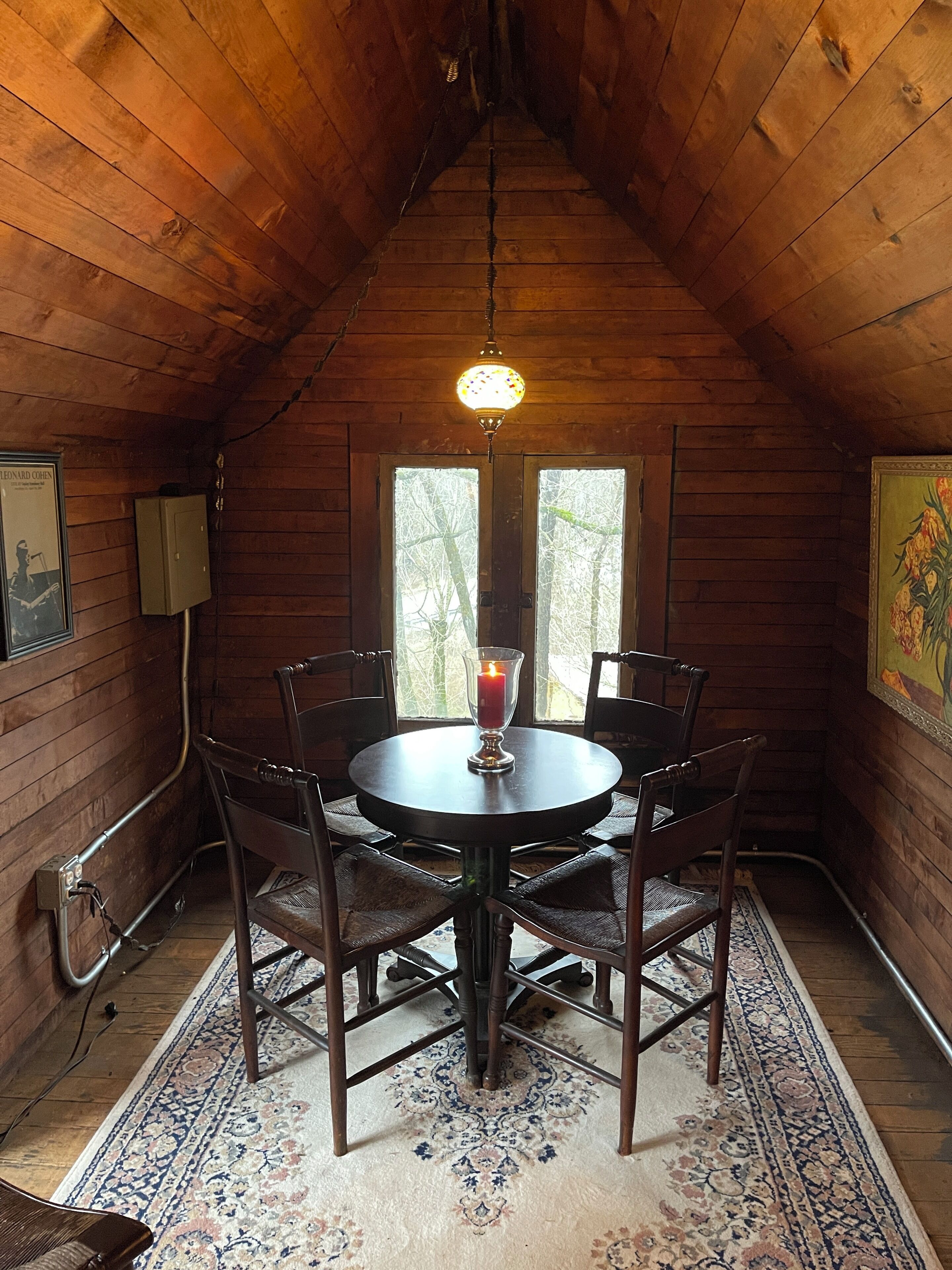 A wooden interior with a round table and chairs, a lit candle, and a patterned rug.