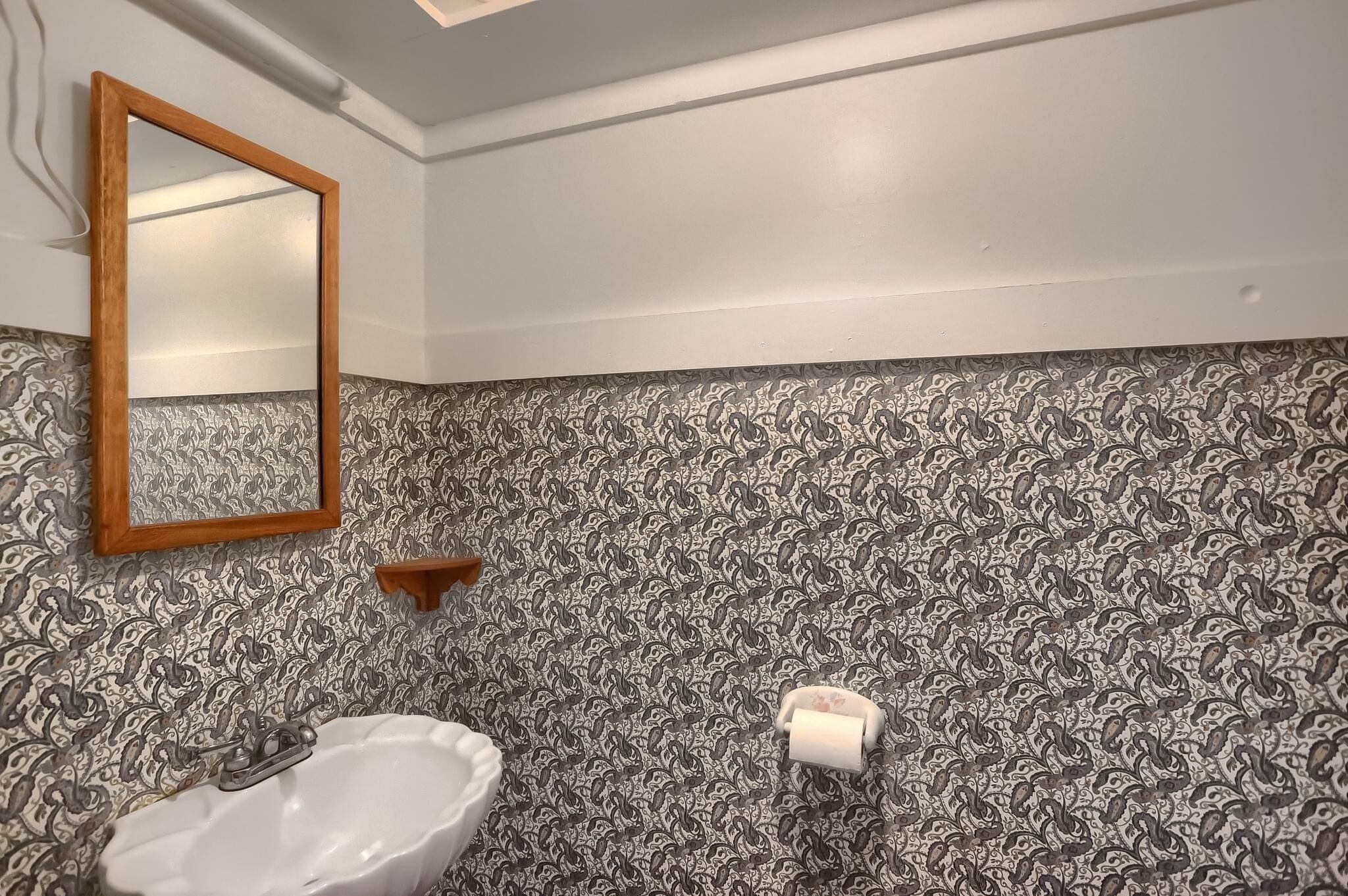 Un baño con pared estampada, espejo con marco de madera, lavabo blanco y una estantería pequeña.