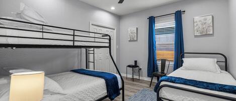 A room with bunk beds, a ceiling fan, a small table with a lamp, and a window with curtains.