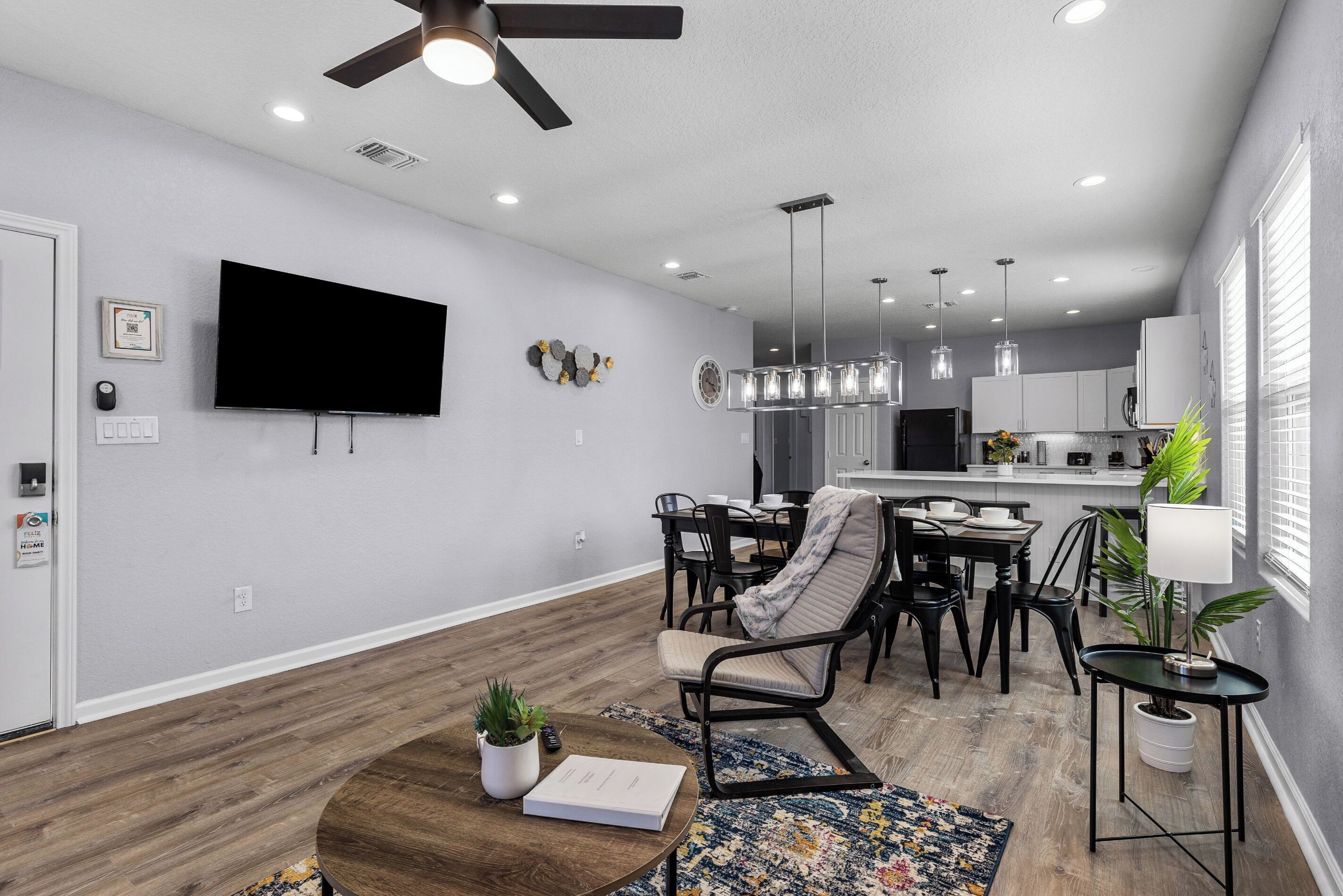 Un salon moderne doté d’un téléviseur à écran plat, d’un ventilateur de plafond, d’un coin repas avec une table et des chaises, et d’une cuisine en arrière-plan.