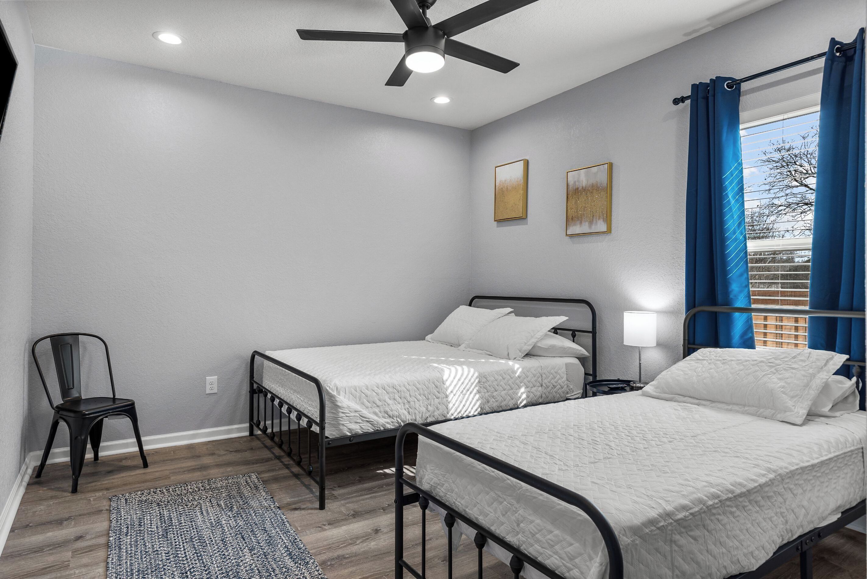 Un dormitorio con dos camas, un ventilador de techo, una silla y una ventana con cortinas.