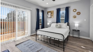 Une chambre à coucher avec un lit, un ventilateur de plafond, une fenêtre avec des rideaux, une lampe fixée au mur et une vue sur l’extérieur.