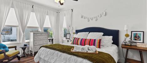 Une chambre à coucher comprenant un lit, un ventilateur de plafond, une commode, une chaise, une table de chevet et une fenêtre avec des rideaux.