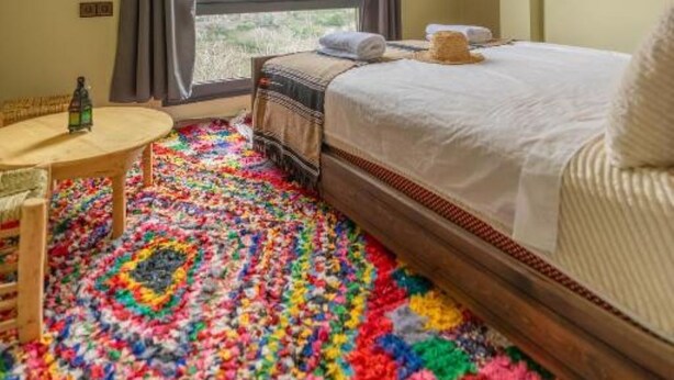 Un dormitori amb un llit gran, una finestra amb cortines, una catifa colorida i una taula petita amb una ampolla.