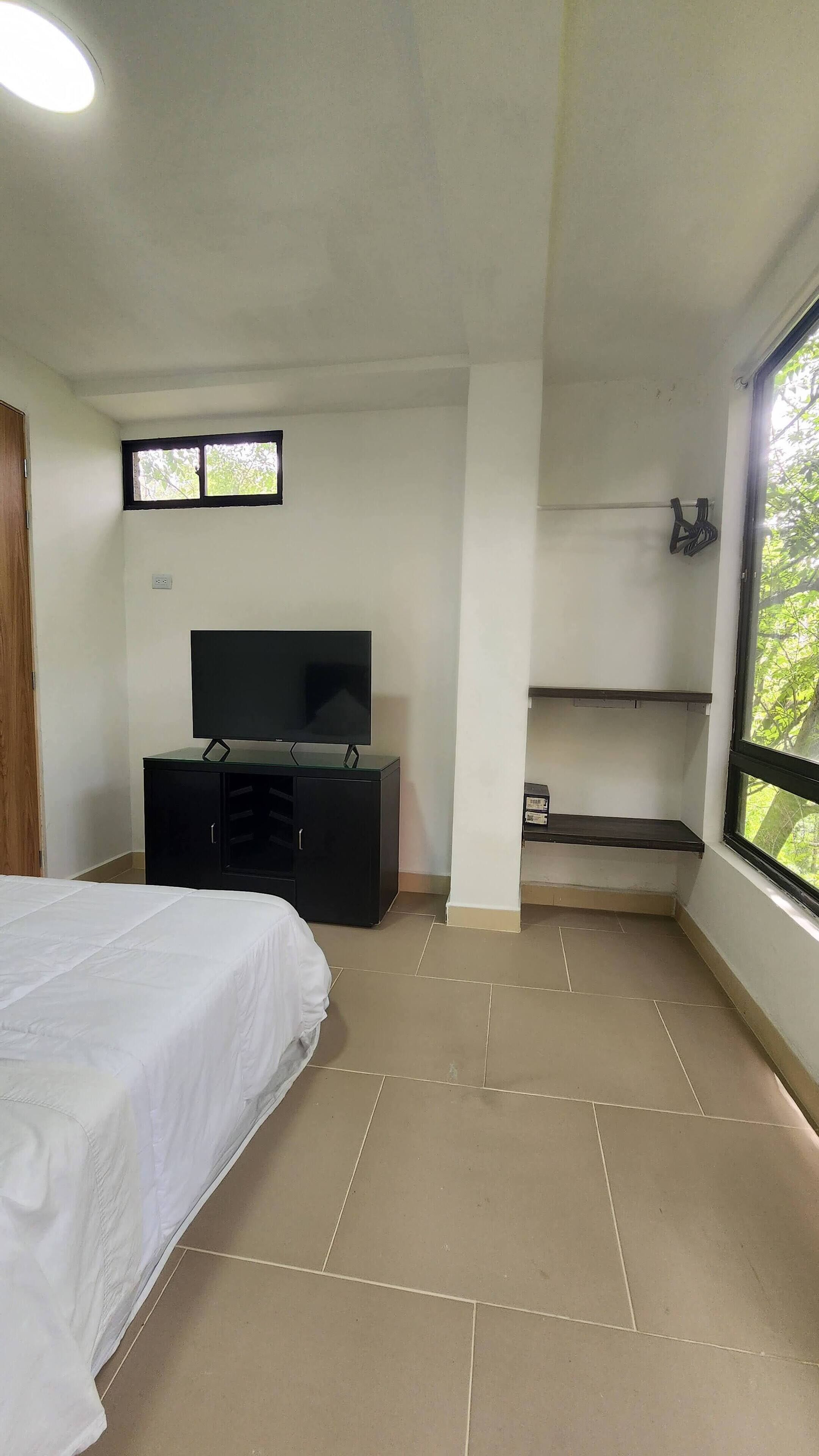 Une chambre à coucher avec un lit, un téléviseur posé sur un meuble noir, une fenêtre et un balcon donnant sur des arbres.