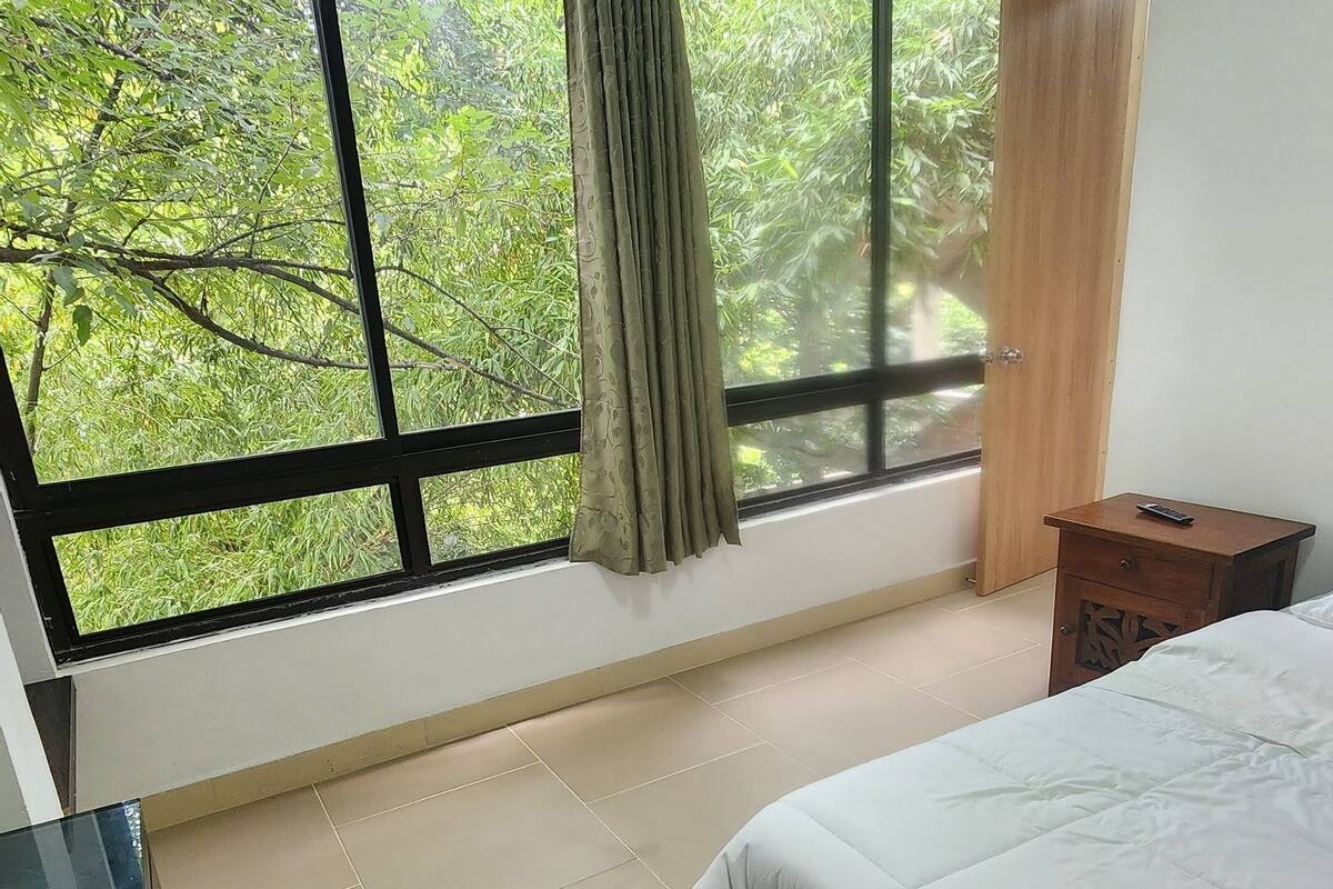 A bedroom with a large window, a bed, a bedside table, and a TV.