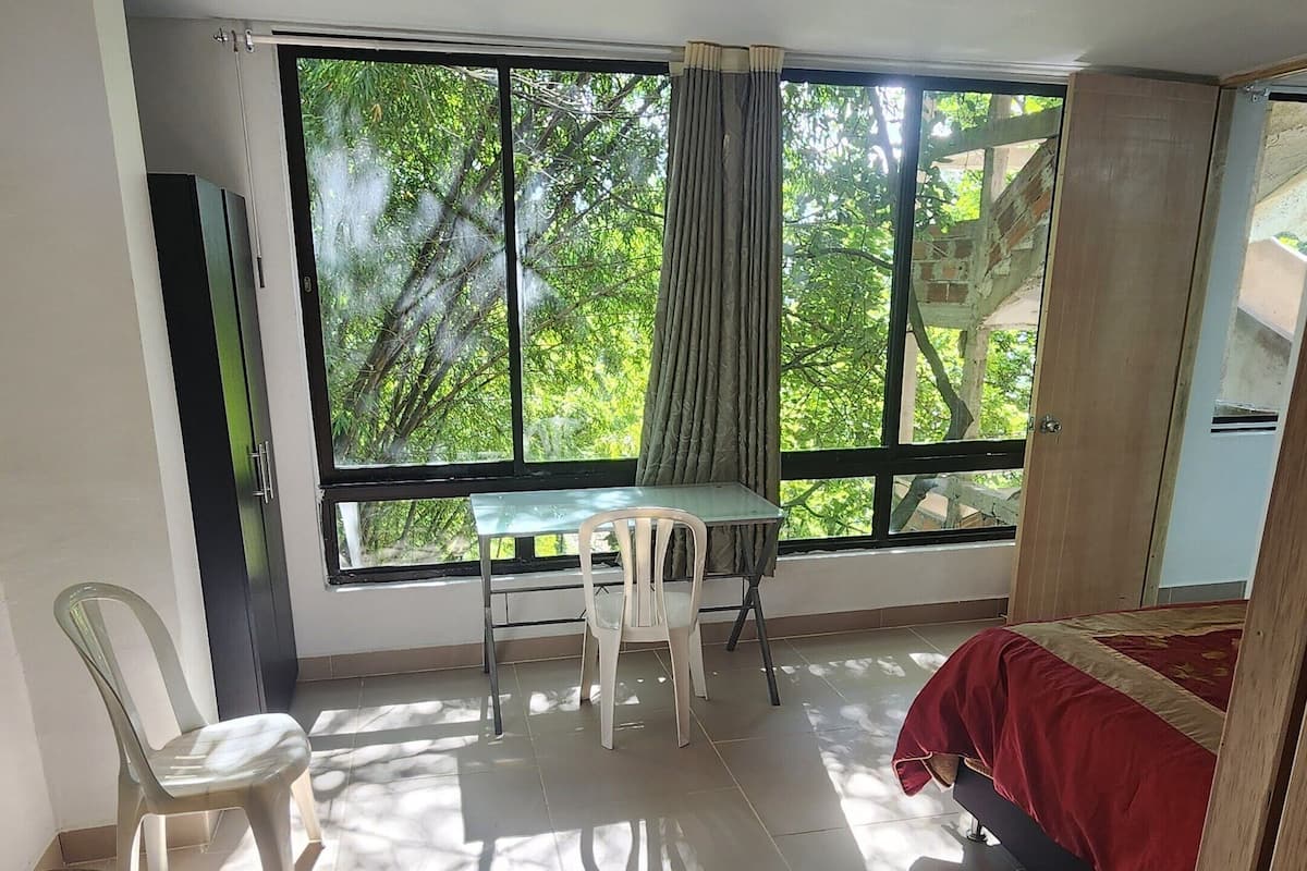 A room with a large window, a table with a chair, a bed, and a wardrobe.