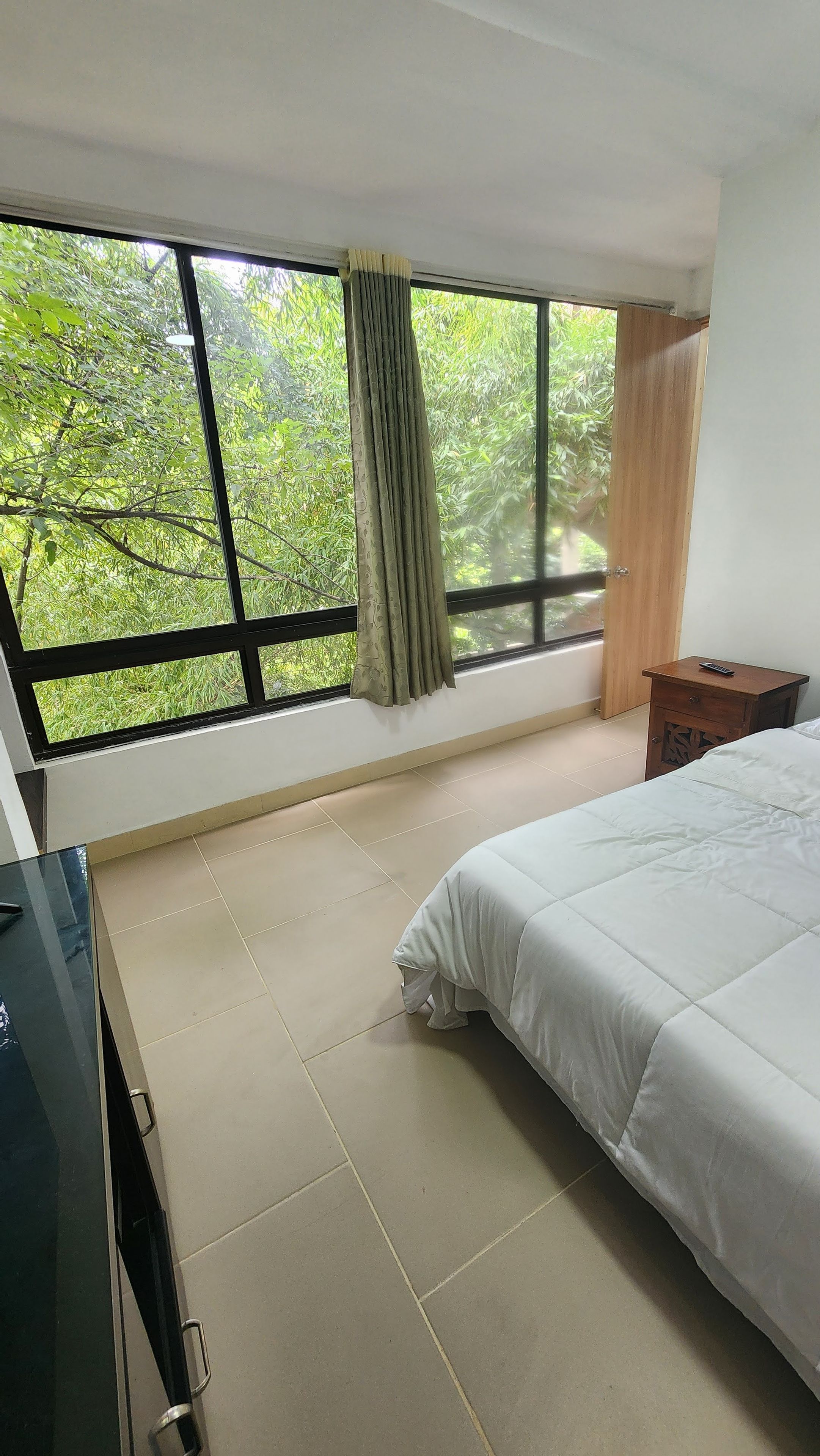 A bedroom with a large window, a bed, a bedside table, and a TV.