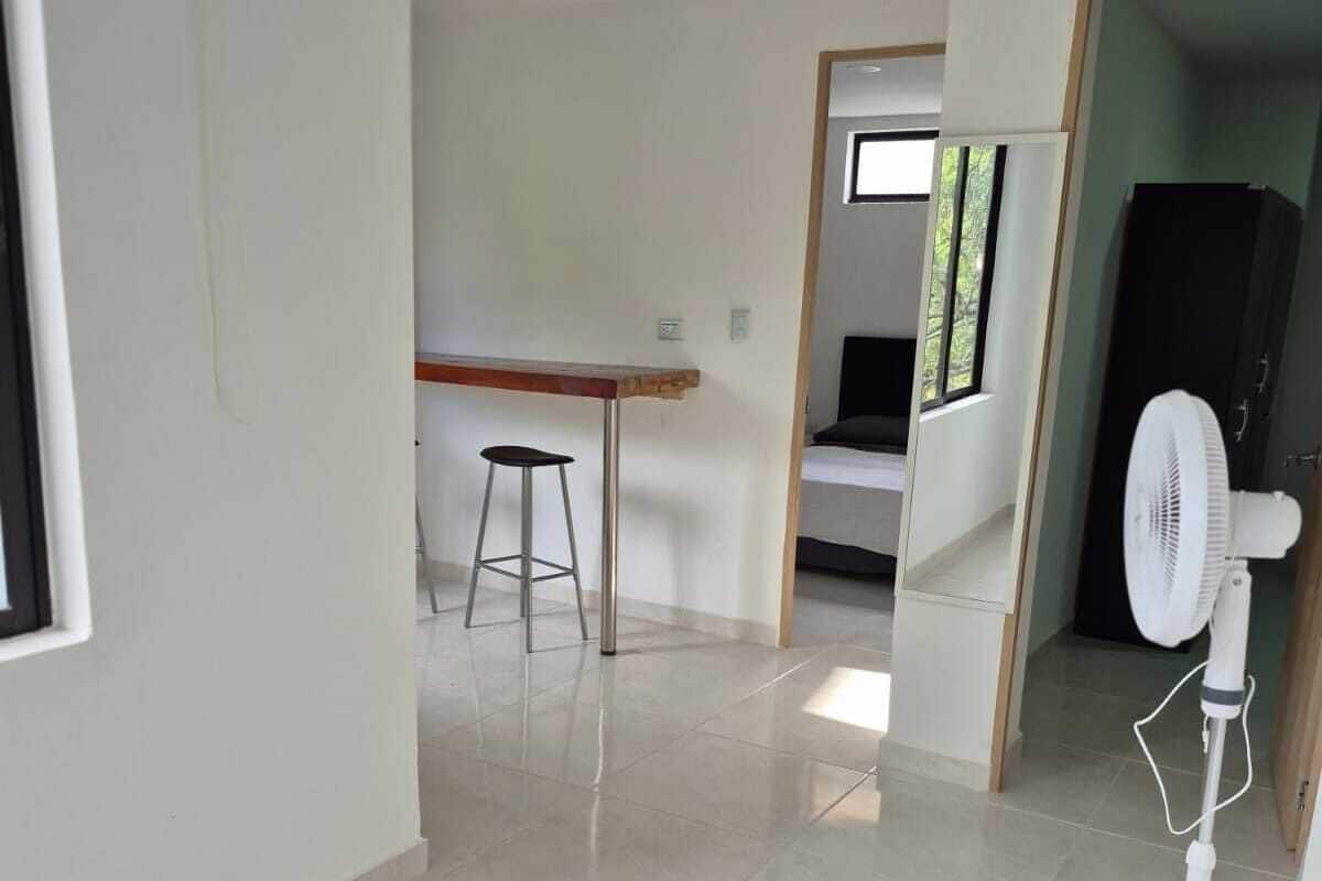 A spacious room with a tiled floor, a standing fan, a desk with a stool, and a bed with a window.