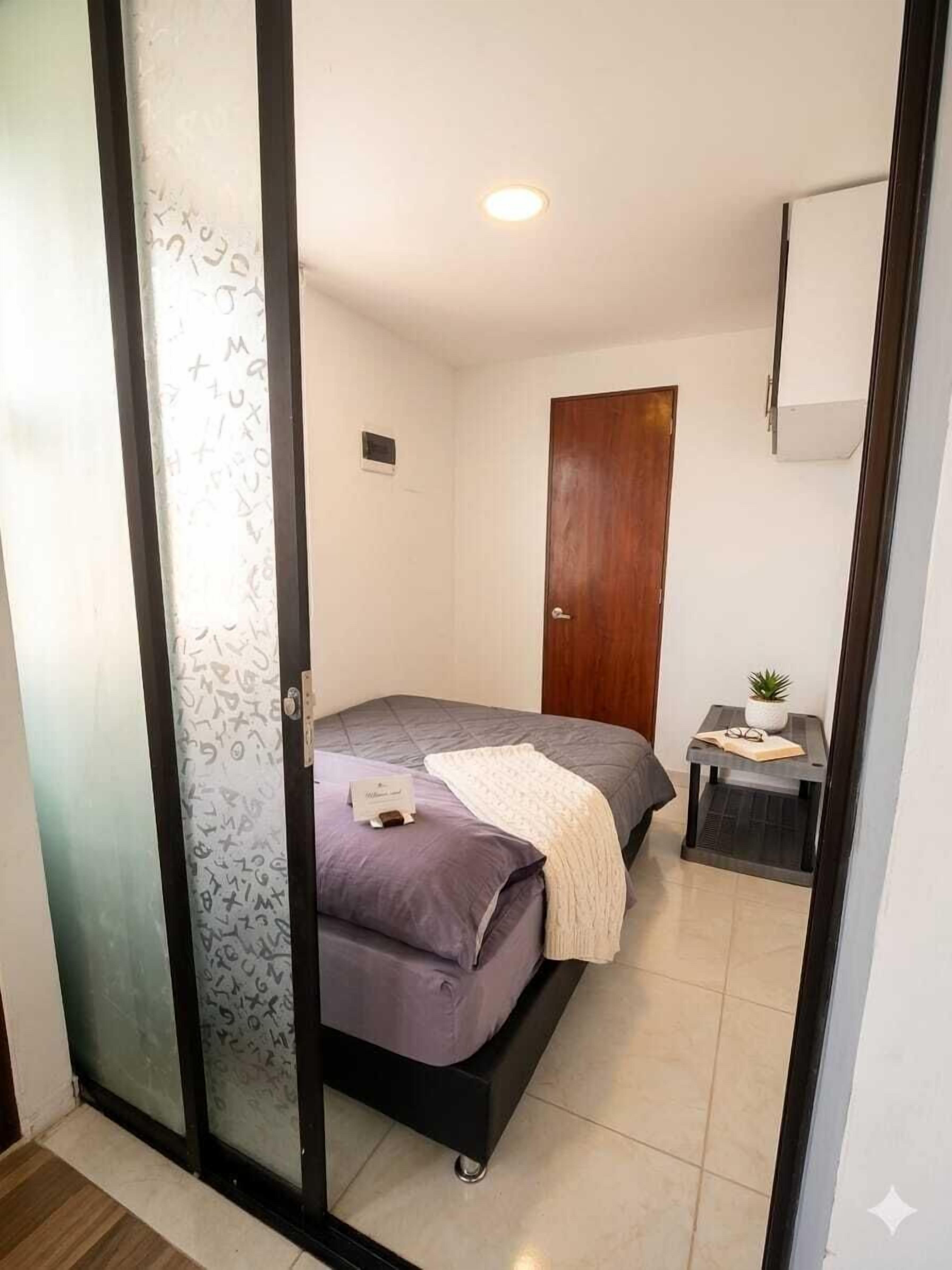 A bedroom with a bed, a wooden door, a small table with a plant, and a wall-mounted air conditioning unit.