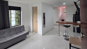 A modern living room with a grey sofa, a wooden door, and a kitchen area with a bar stool and a refrigerator. - PILARICA GARDENS (Medellín)
