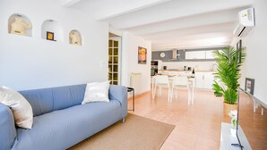 Uma sala de estar moderna com um sofá azul, uma área de jantar com uma mesa e cadeiras brancas, e uma cozinha ao fundo.