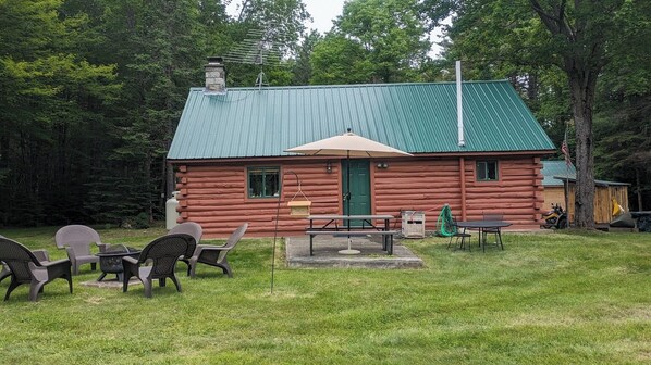 A log cabin with a green metal roof, a green door, and a small porch area with outdoor seating.