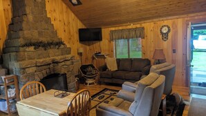 L’intérieur chaleureux d’un chalet avec une cheminée en pierre, des murs en bois, un téléviseur à écran plat, un canapé et un coin repas avec des chaises.
