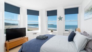 A bedroom with a large bed, a TV on a wooden stand, and multiple windows offering ocean views. - 3 bedroom accommodation in Woolacombe (Woolacombe)