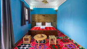 Een kamer met een blauwe wand, een hoofdbord met baksteenmotief, een bed met witte kussens, een vloerkleed met rood-zwarte patronen, een houten tafel met een hoed, en twee kleine houten krukjes.