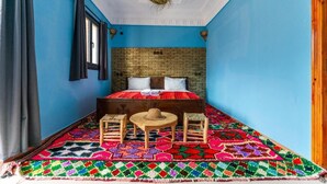 Een kamer met een blauwe wand, een hoofdeinde met baksteenpatroon, een kleurrijk geometrisch vloerkleed, een klein rond tafeltje, twee houten krukken en een raam met gordijnen.