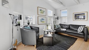 Ett modernt vardagsrum med en soffa i läder, en svart fåtölj och ett soffbord. På väggarna hänger flera inramade konstverk, och det finns även en krukväxt och en golvlampa.
