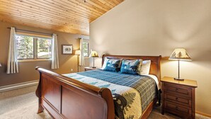 A bedroom with a wooden ceiling, a bed with a patterned bedspread, two bedside lamps, and a nightstand with drawers. - Woodbine Cabin | Big Sky (Big Sky)