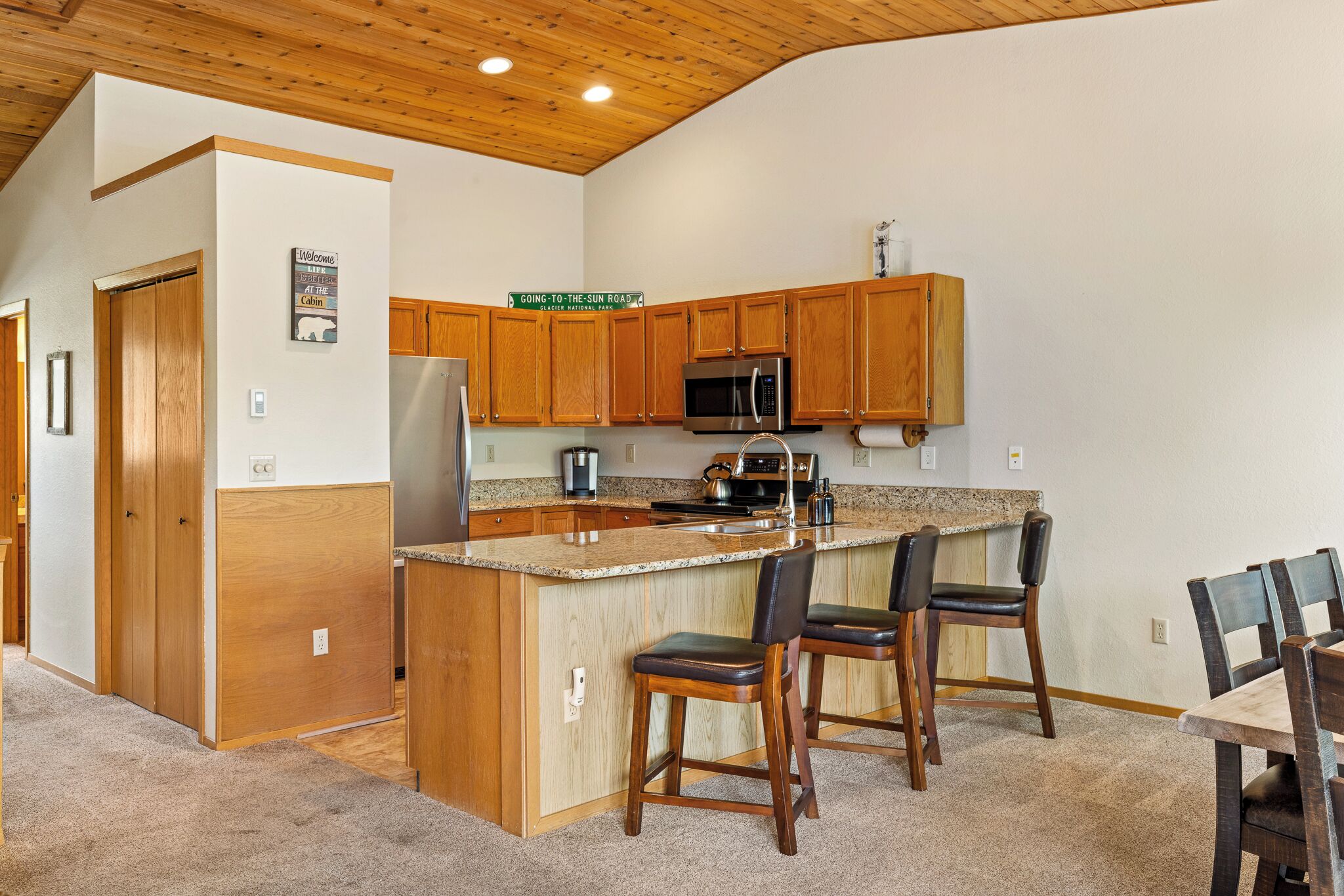 A kitchen with wooden cabinets, a stainless steel refrigerator, a microwave, and a granite countertop. There are two bar stools and a dining table with chairs.