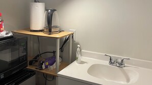 Ett badrum med ett vitt handfat, en spegel, en hållare för pappershanddukar, en mikrovågsugn och en skrivbordslampa.