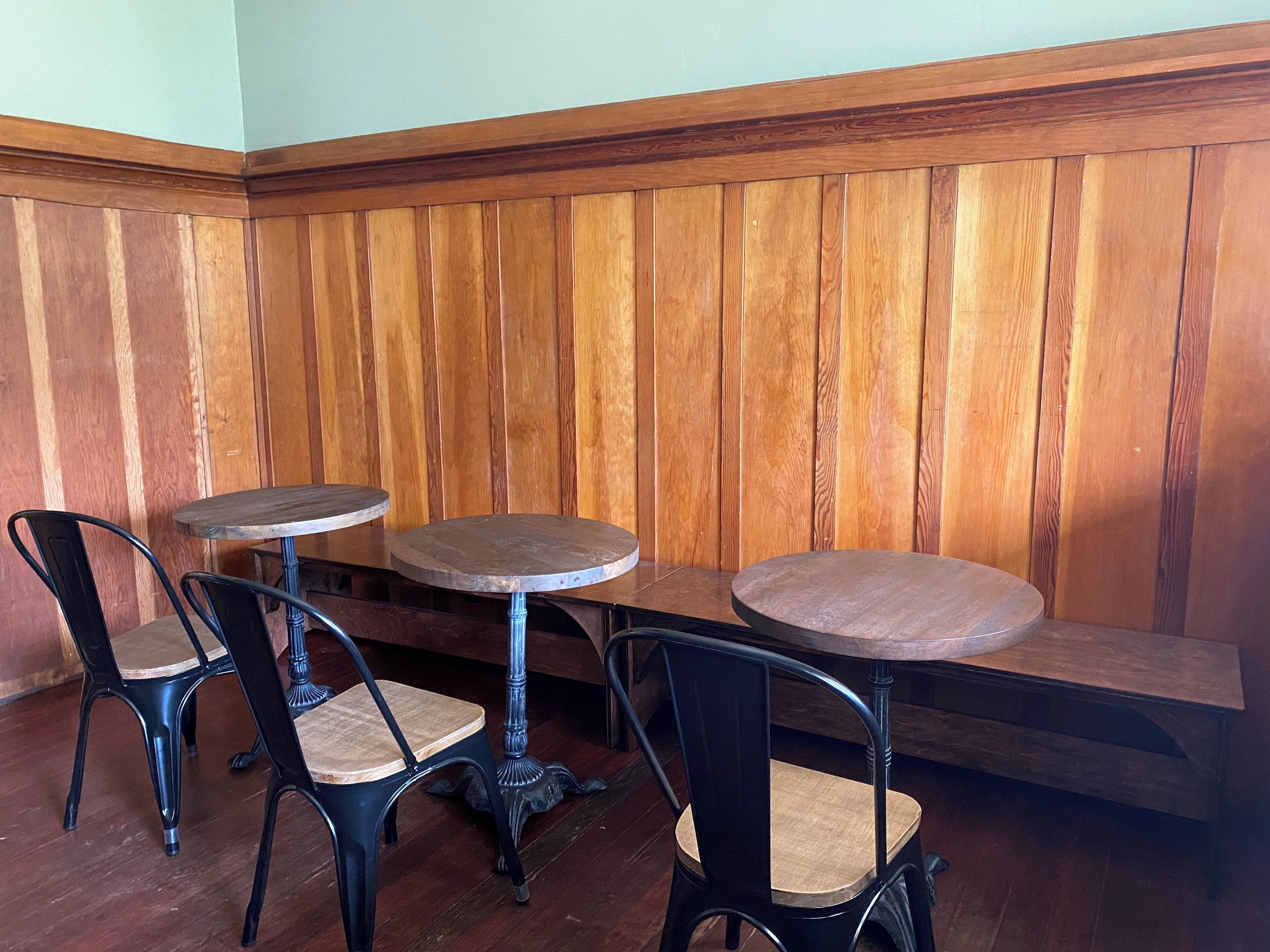 A corner seating area with wooden paneling, two round tables, and four chairs with metal frames and cushioned seats.