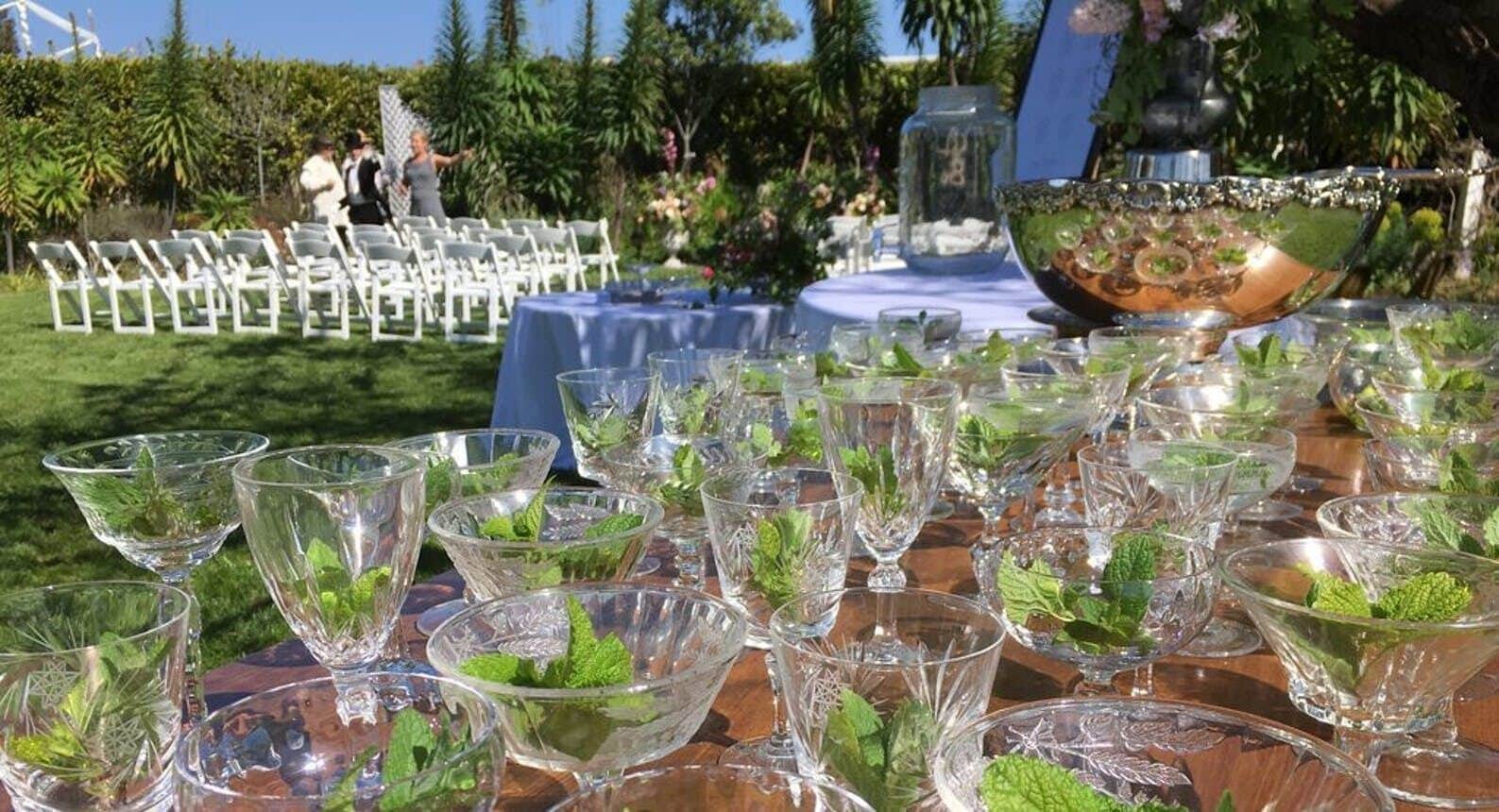 A table set outdoors with clear glassware and mint leaves, surrounded by white chairs and a blue tablecloth.