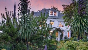 A large, two-story house with a symmetrical facade, surrounded by lush greenery and flowering plants. - The Weller House (Fort Bragg)