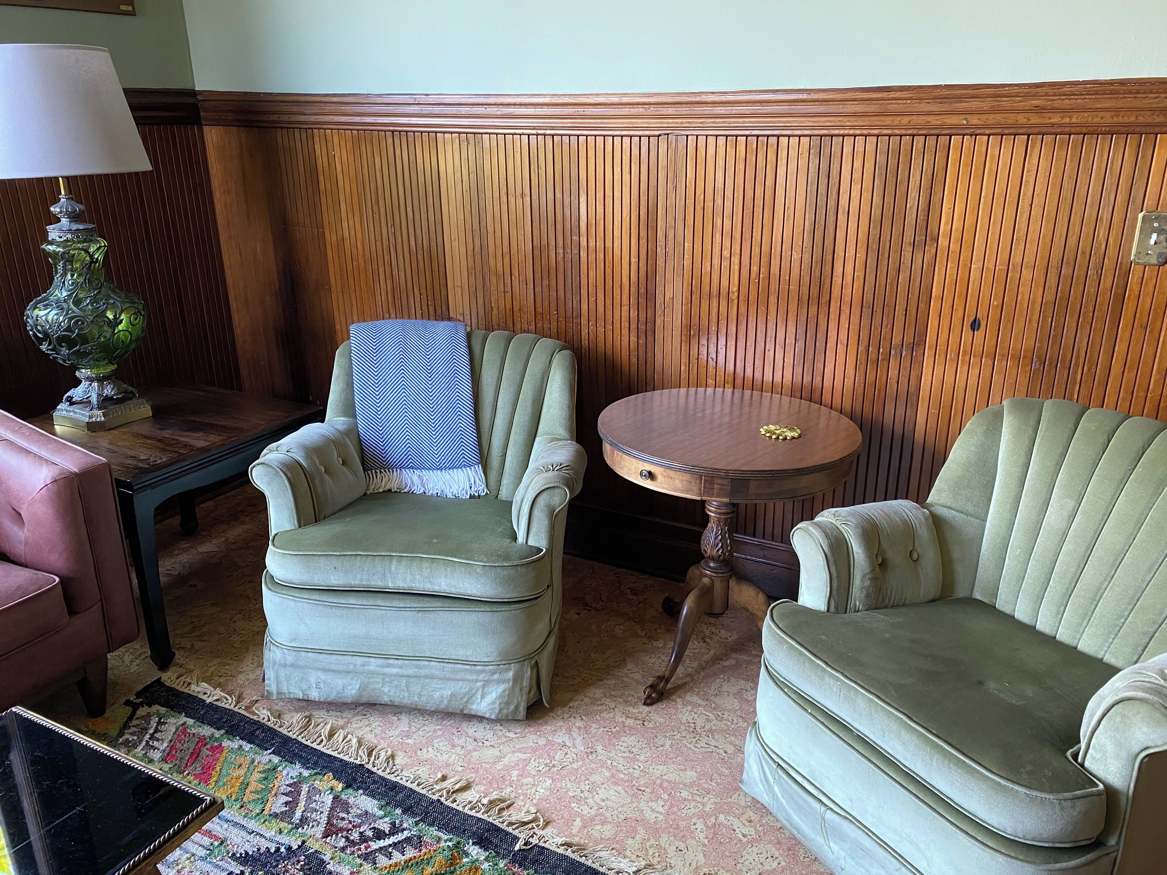 Une pièce avec un mur lambrissé en bois, un fauteuil vert, un canapé assorti, une petite table ronde et une lampe.