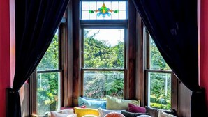 A cozy room with a window seat adorned with colorful cushions, a stained glass window, and dark curtains. - The Weller House (Fort Bragg)