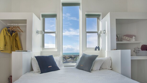 A bedroom with a large bed, two windows, a closet, and a view of the sea. - The Weller House (Fort Bragg)