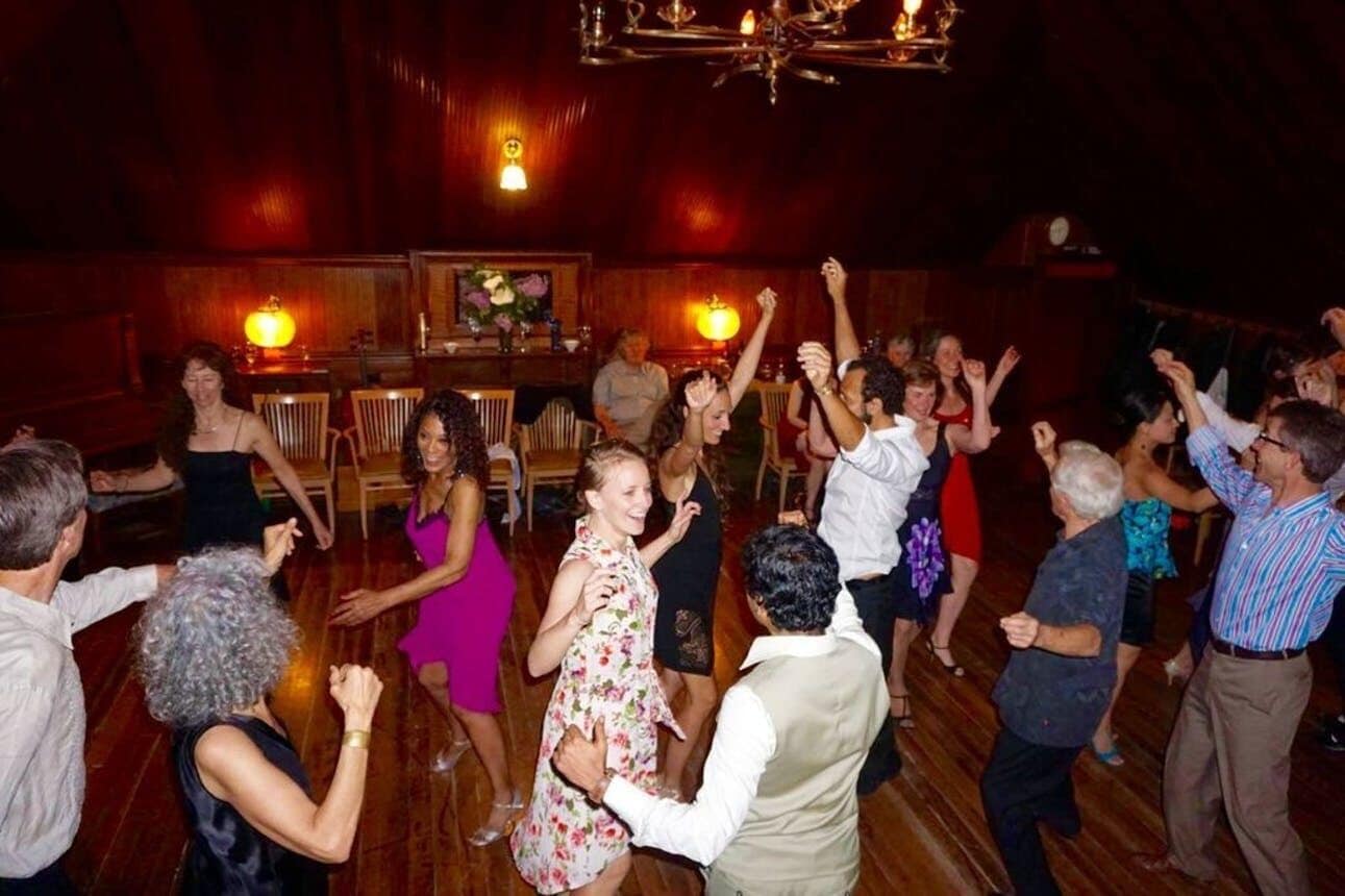 Un grupo de personas bailando en una sala tenuemente iluminada, con pisos de madera y un candelabro.