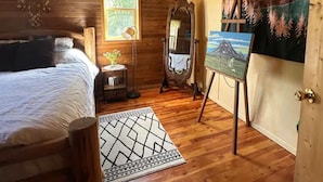 A cozy wooden cabin bedroom with a bed, a dresser, a mirror, and a painting on the wall. - Cedar Cabin Cozy 2-Bedroom next to Glacier Park (East Glacier Park)