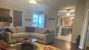 A living room with a sectional sofa, a coffee table, and a floral rug. There is a kitchen in the background with a dining table and chairs.