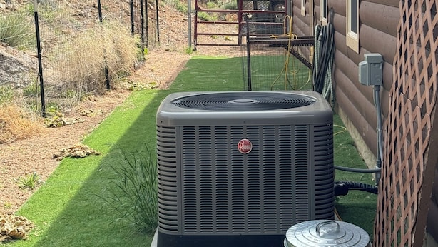 An outdoor air conditioning unit in front of a wooden house with a metal gate and a grassy lawn.