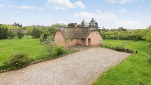Een rieten huis met een grindpad, omgeven door weelderige groene velden en bomen.