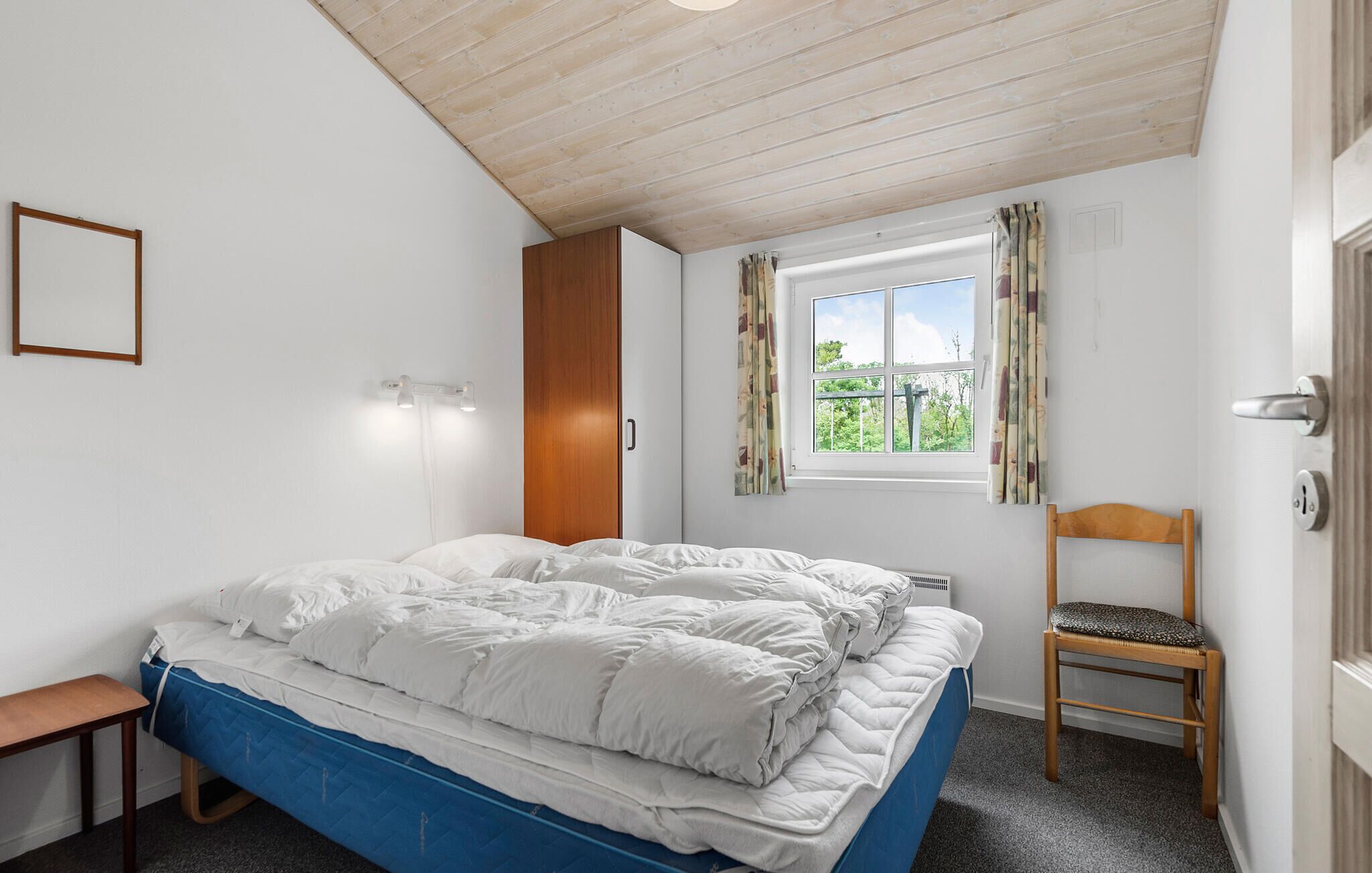 A bedroom with a bed, a chair, a window with curtains, a wooden ceiling, and a door.