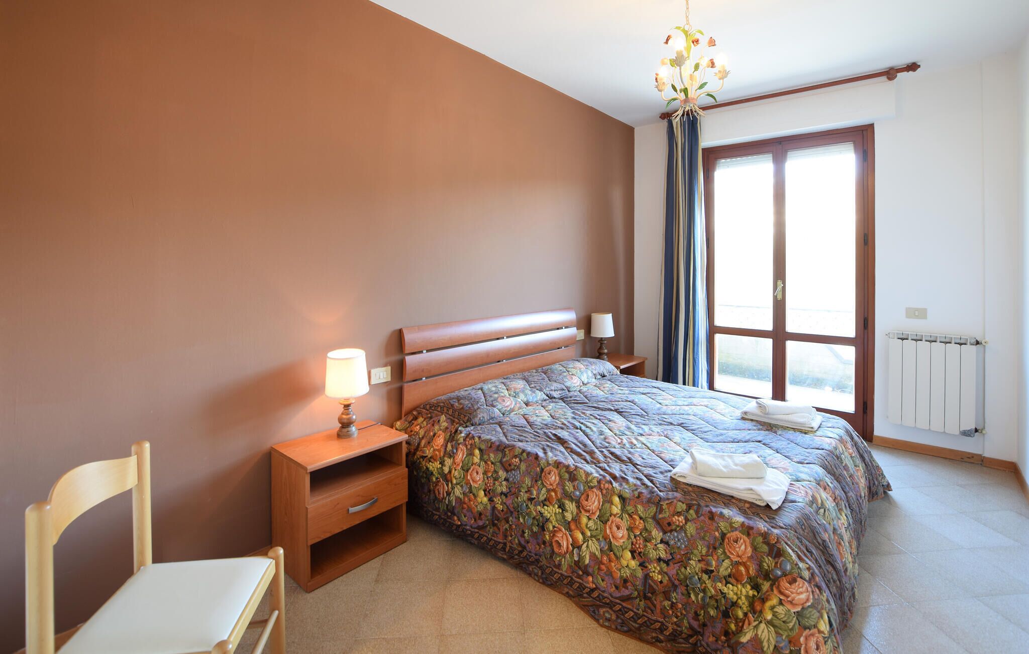 Un dormitorio con cama, mesita de noche, silla y ventana con cortinas.