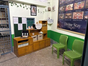 Un área de recepción con un escritorio de madera, sillas verdes, una pared con el letrero 'Divine Stay' y un cartel con varios nombres.