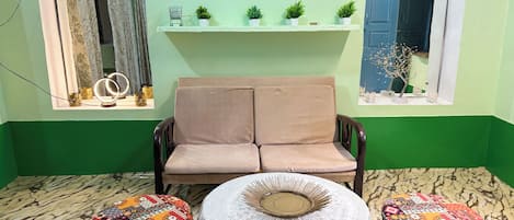 A room with a sofa, two patterned ottomans, a small table with a lace tablecloth, and a shelf with plants and a clock.