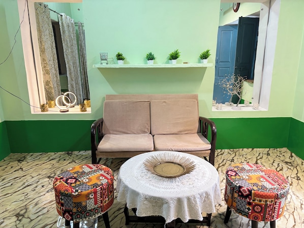 A room with a sofa, two patterned ottomans, a small table with a lace tablecloth, and a shelf with plants and a clock.