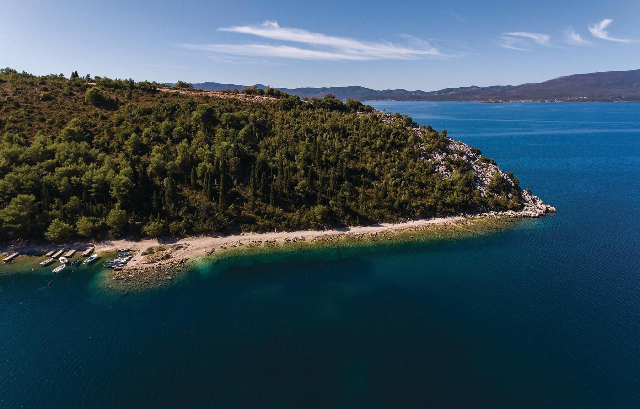 An aerial view of a coastal area with a forested peninsula, clear blue waters, and boats docked along the shore.
