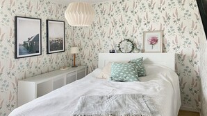 A bedroom with floral wallpaper, a bed with a white and grey blanket, two framed pictures on the wall, a bedside table with a lamp, and a small decorative wreath. - Stunning home in Gotland with kitchen (Gotland)