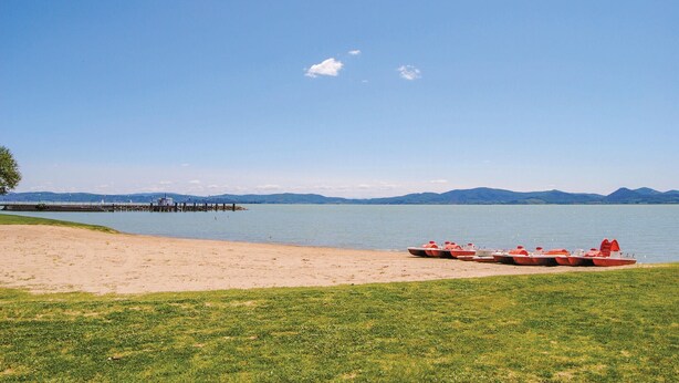 A beach with a pier extending into calm water, a grassy area, and mountains in the background.