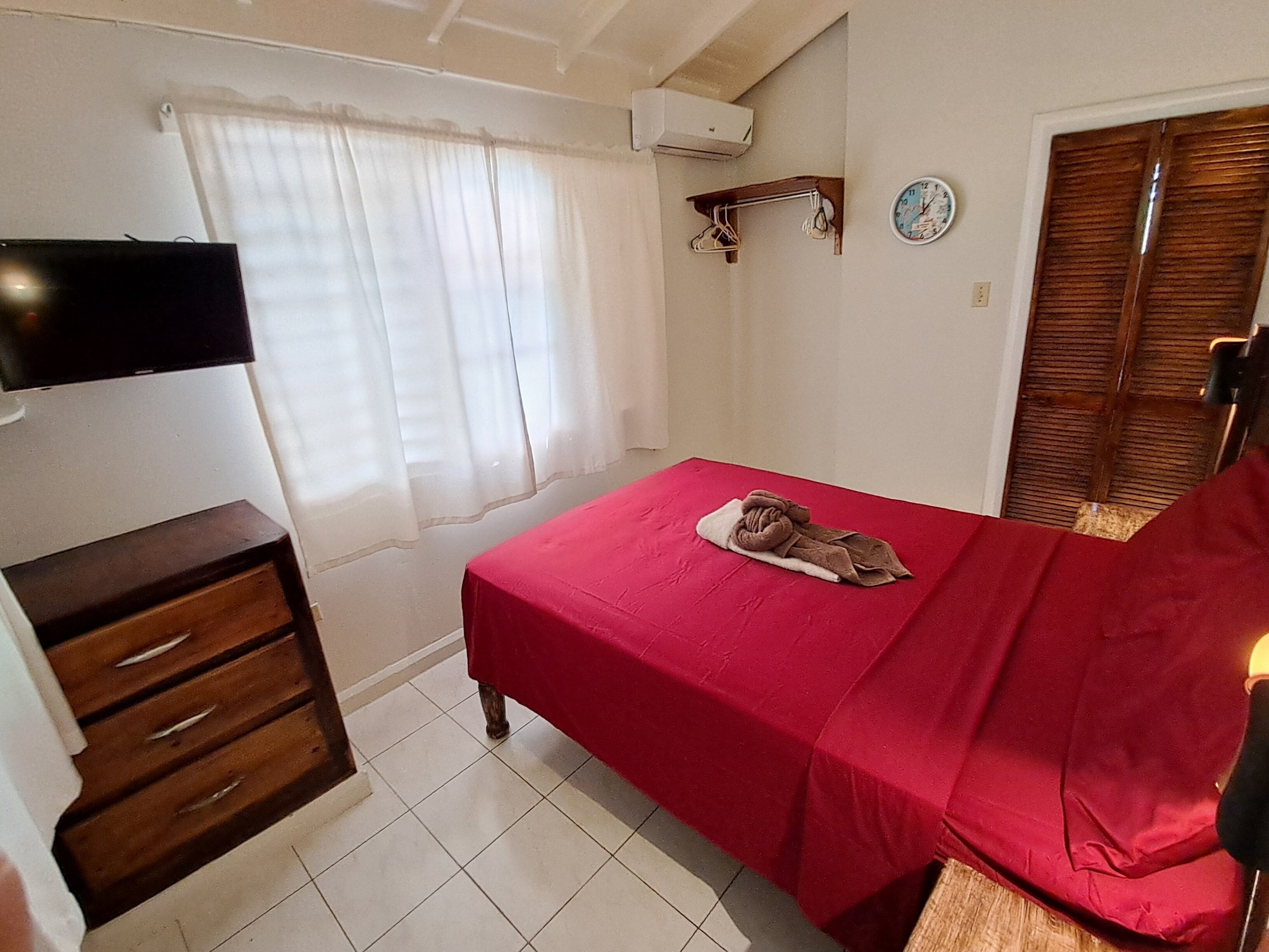 Una habitación pequeña y ordenada con una cama, un televisor montado en la pared, un aparador de madera, una ventana con cortinas y un reloj en la pared.