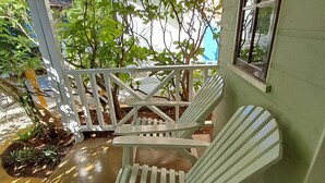 A covered porch with two white Adirondack chairs, a concrete floor, and a view of a garden and street. - Barry's Rooms Negril (Negril)
